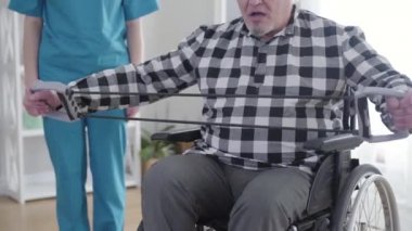 Tekerlekli sandalyedeki yaşlı gri saçlı beyaz adam arka planda tanınmayan profesyonel hemşire olarak egzersiz yapıyor. Esnek lastikleri esneten eski engelli emekliler. Engelli, rehabilitasyon.