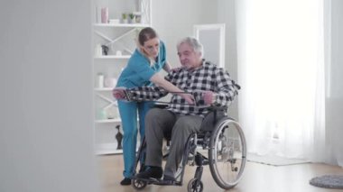 Kafkasyalı bir kadının yaşlı adama elastik bant takmasına yardım ettiği uzun bir çekim. Olgun emekliler huzurevinde yatalak ihalenin yardımıyla egzersiz yapıyorlar. Rehabilitasyon, fizik tedavi.