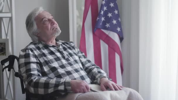 Portrait of upset thoughtful Caucasian man looking away and thinking. Sad grey-haired guy holding army stripe in hands and sitting in wheelchair. Military discharge, veterancy, lifestyle. — Stock video