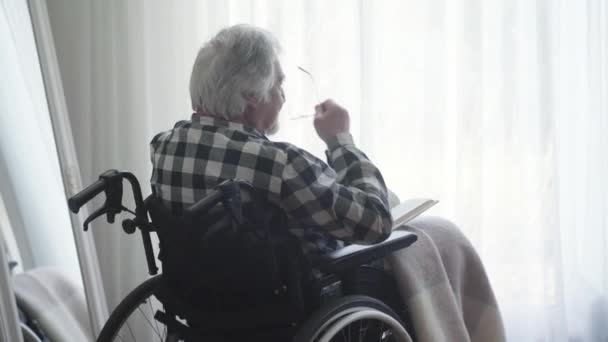 Vista posterior lateral del anciano hombre caucásico poniéndose anteojos y leyendo libro. Vieja jubilada discapacitada sentada en silla de ruedas en casa en un día soleado. Discapacidad, jubilación, problemas médicos . — Vídeos de Stock