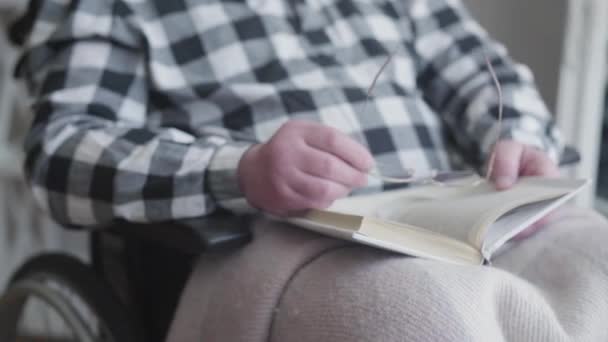 Vieux caucasien méconnaissable tenant livre et lunettes comme assis dans un fauteuil roulant. Retraité handicapé lisant à la maison. hobby, retraite, handicap, mode de vie . — Video