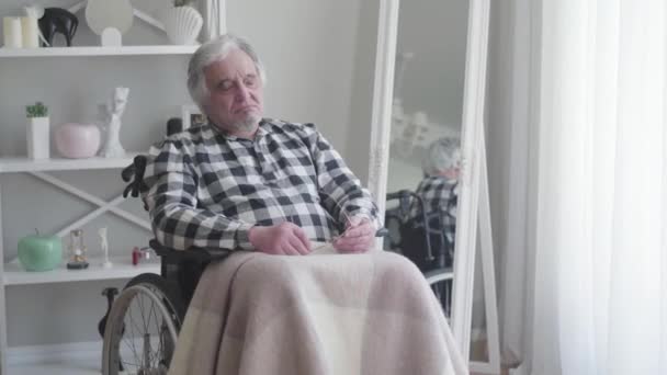 Elderly Caucasian grey-haired man sitting in wheelchair and signing. Old male retiree suffering from severe pain in knee. Aging, oldness, disability, illness. — Stock video