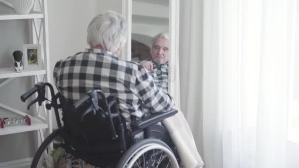 Achteraanzicht van een volwassen blanke man in een rolstoel die naar zijn spiegelbeeld kijkt. Droevige bejaarde gepensioneerde brengt de dag alleen door binnen. Herinneringen, veroudering, pensioen, levensstijl. — Stockvideo