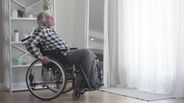 Kafkasyalı, üzgün, engelli bir adamın portresi. Tekerlekli sandalyeyi pencereden indiriyor ve yuvarlanıyor. Depresyondaki erkek emekli, içeride hareket etmek için ekipman kullanıyor. Yalnızlık, sakatlık, yaşlanma.. — Stok video
