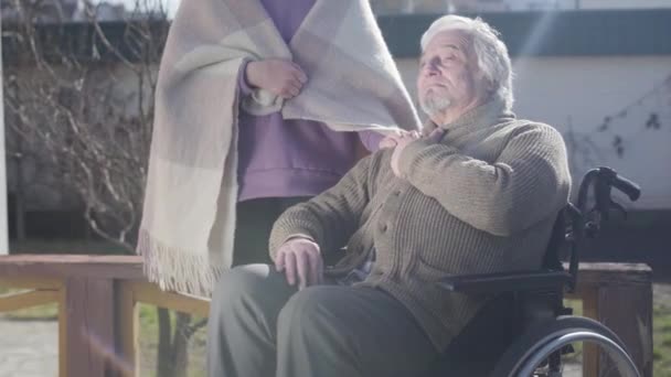 Positive old Caucasian disabled man in wheelchair sitting outdoors in sunrays and talking with unrecognizable nurse. Invalid tender taking care of elderly retiree. Disability, lifestyle, assistance. — Stock video