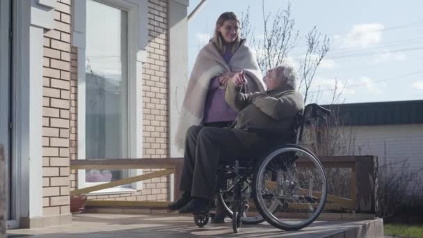 Dorosła kaukaska pielęgniarka w okularach rozmawiająca z niepełnosprawnym staruszkiem na wózku inwalidzkim i uśmiechnięta. Nieprawidłowy przetarg spędzający słoneczny dzień z pacjentem na świeżym powietrzu. Pomoc, rehabilitacja, niepełnosprawność. — Wideo stockowe
