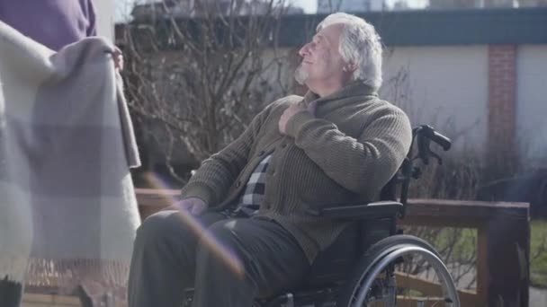 Genç, tanınmayan bir kadın, kötürüm, yaşlı, beyaz bir adamın battaniyeli bacaklarını kapatıyor. Tekerlekli sandalyedeki mutlu erkek emekli, güneşli bir günü dışarıda hemşireyle geçiriyor. Yardım, rehabilitasyon. — Stok video