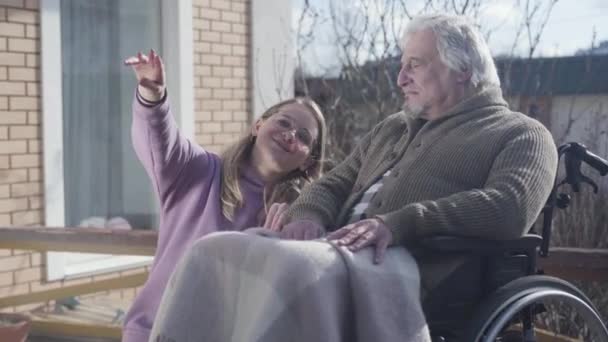 Pozytywna biała kobieta w okularach siedząca obok starego niepełnosprawnego mężczyzny na wózku inwalidzkim i pokazująca coś. Profesjonalna pielęgniarka zabawiająca emeryta na świeżym powietrzu. Dom opieki, pomoc, pomoc. — Wideo stockowe