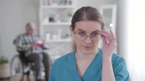 Close-up portrait of confident adult Caucasian woman in eyeglasses looking at camera and smiling. Professional invalid tender gesturing yes by shaking head. Blurred disabled man at the background. — Stock Video