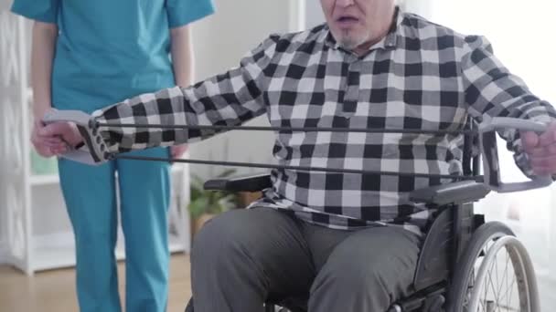 Äldre gråhårig vit man i rullstolsträning som professionell oigenkännlig sjuksköterska stående vid bakgrunden. Gamla handikappade pensionär sträcker elastiskt band. Funktionshinder, rehabilitering. — Stockvideo