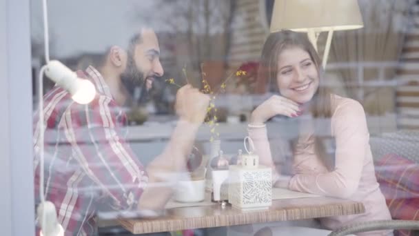 Strzelanie przez szybę w oknie młodej wieloetnicznej pary zakochanej siedzącej przy stole w kawiarni i rozmawiającej. Uśmiechnięty mężczyzna z Bliskiego Wschodu i biała kobieta spotykająca się w restauracji. Miłość, styl życia, radość. — Wideo stockowe