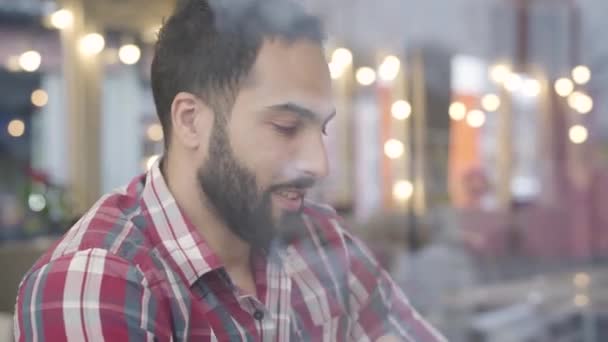 Retrato de primer plano del guapo hombre de Oriente Medio sentado detrás de un cristal de ventana en la cafetería y hablando con una persona desconocida. Joven barbudo chico citas con novia o esposa en el restaurante. Ocio, estilo de vida — Vídeos de Stock