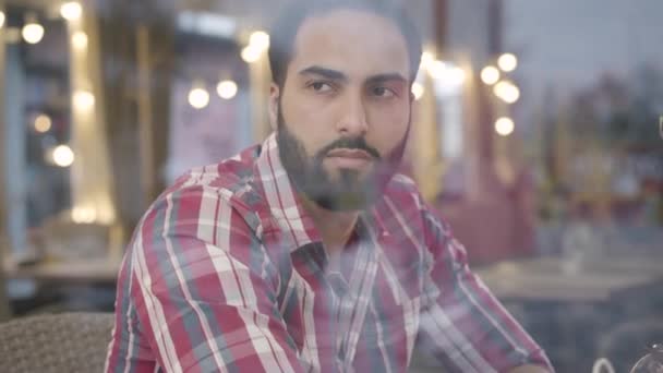 Πυροβολώντας μέσα από τζάμι παραθύρου ενός όμορφου άντρα από τη Μέση Ανατολή που κάθεται στην καφετέρια και μιλάει. Πορτρέτο του νεαρού ανήσυχου άντρα με μαύρα μαλλιά και καστανά μάτια. Ανησυχίες, προβλήματα, τρόπος ζωής. — Αρχείο Βίντεο