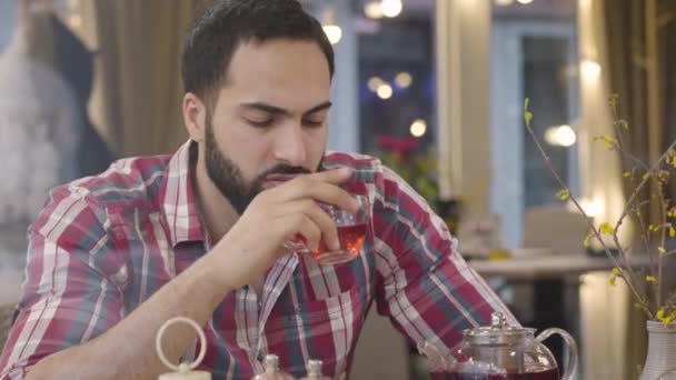 Un homme du Moyen-Orient stressé buvant du thé chaud dans un café, regardant ailleurs, secouant la tête et signant. Portrait d'inquiet jeune beau garçon au restaurant réfléchissant à la solution du problème. Anxiété, stress . — Video