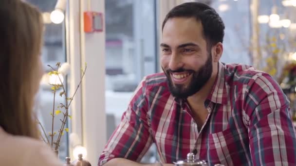 Retrato de homem barbudo positivo do Oriente Médio rindo em voz alta de piadas de sua namorada caucasiana irreconhecível. Casal multi-étnico jovem namoro no café e conversando. Alegria, estilo de vida, felicidade . — Vídeo de Stock