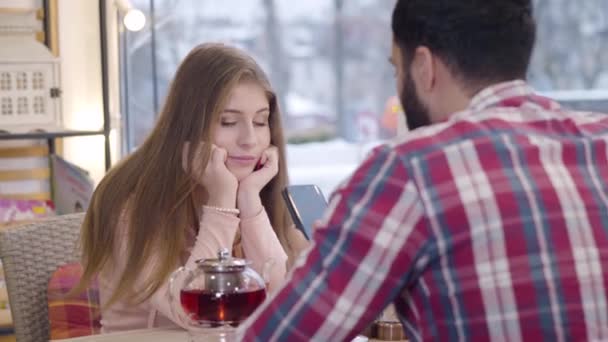 Retrato de menina bonita caucasiana entediado sentado com namorado ou marido no restaurante como seu cônjuge usando smartphone. Homem barbudo do Médio Oriente ignorando o belo parceiro. Namoro, decepção . — Vídeo de Stock