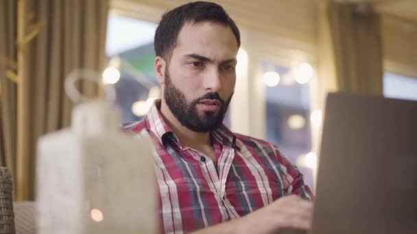 Detailní portrét koncentrovaného muže ze Středního východu, který sedí u stolu v kavárně a píše na klávesnici notebooku. Důvěryhodný podnikatel pracující online v restauraci. Životní styl, přepracování, obchod. — Stock video