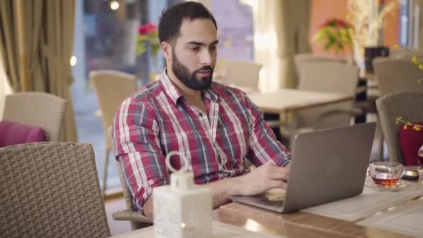 Plan médian d'un homme d'affaires confiant du Moyen-Orient travaillant sur un ordinateur portable dans un restaurant. Concentré jeune homme tapant sur le clavier comme assis à la table dans le café. Mode de vie, affaires, entrepreneuriat . — Video