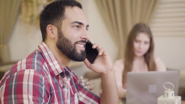 Погляд на усміхненого бізнесмена з Близького Сходу, який розмовляє по телефону, як розмита молода кавказька жінка, що працює на ноутбуці на задньому плані. Підприємці під час обіду працюють у кафе.. — стокове відео