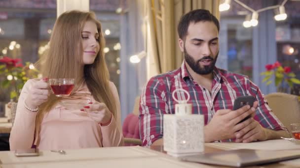 Retrato de una linda chica caucásica mirando al novio usando un teléfono inteligente y riendo. Hombre de Oriente Medio escondiendo el teléfono y sonriendo. Joven pareja alegre descansando en la cafetería. Relación, alegría, citas . — Vídeos de Stock