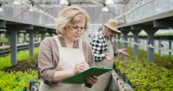 文書中の植物の成長を示す眼鏡でプロの白人女性農学者の肖像画。温室に立って周りを見ている快適な女性。シネマ4k｜ProRes HQ. — ストック動画