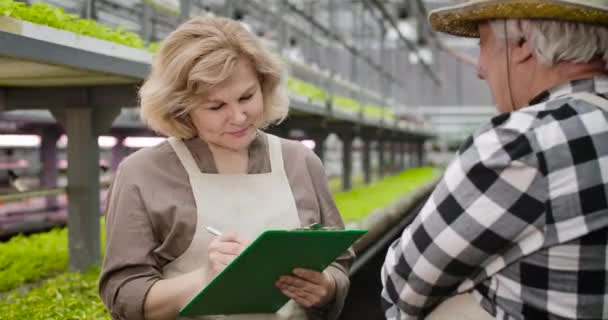 Glada äldre blonda kaukasiska kvinna med grå ögon lyssna på mogna manliga agronomen i växthus och skriva information om grönsaker tillväxt. Jordbruk, odling. Cinema 4k Prores Hq. — Stockvideo