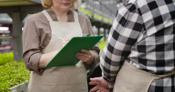 Unerkennbare kaukasische reife Frau, die mit Mann im Glashaus spricht und Informationen schreibt. Ernsthafte professionelle Agronomen, die Daten über das Gemüsewachstum sammeln. Cinema 4k ProRes Hauptquartier. — Stockvideo