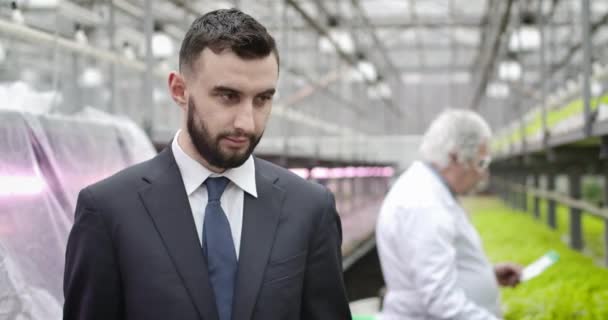 Retrato de homem branco bonito jovem com cabelo preto e olhos castanhos posando em estufa. Empresário confiante de terno olhando para a câmera como biólogo trabalhando em segundo plano. Cinema4k ProRes HQ — Vídeo de Stock