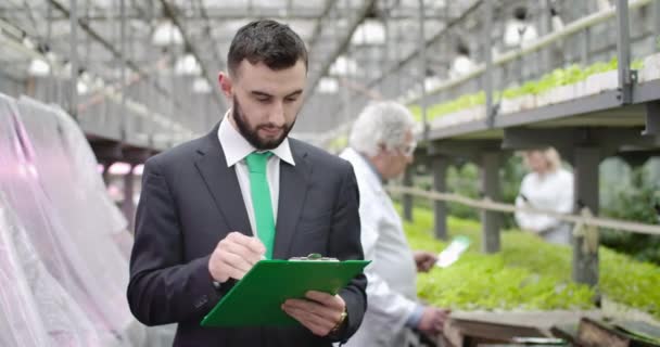 Retrato de um jovem caucasiano confiante a inspeccionar uma estufa. Bonito auditor barbudo tomando notas e olhando para a câmera. Agricultura, controle, negócios, produção. Cinema 4k ProRes HQ . — Vídeo de Stock