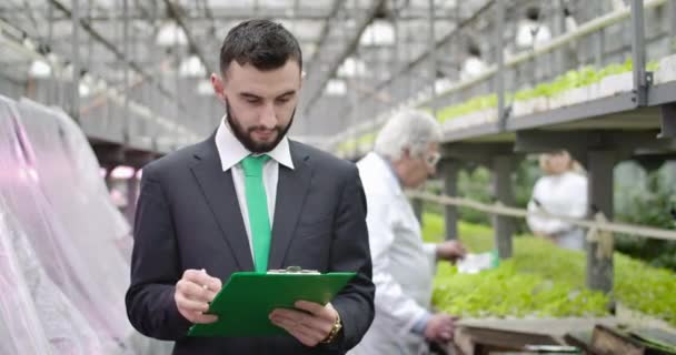 Junger kaukasischer bärtiger Mann, der einen reifen, verschwommenen Agronomen fragt und sich umdreht, um Informationen zu schreiben. Zuverlässiger Prüfer, der in einem Gewächshaus arbeitet, das Bio-Gemüse anbaut. Cinema 4k ProRes Hauptquartier. — Stockvideo