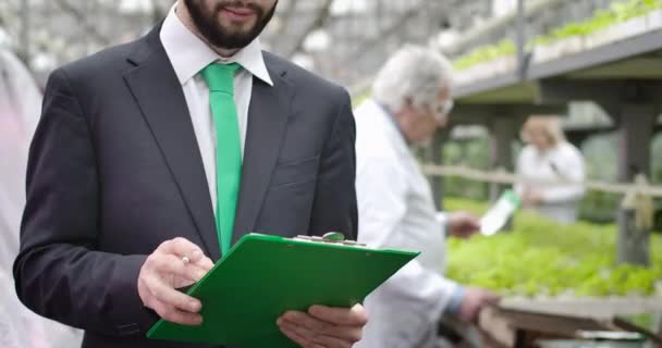 Onherkenbare blanke jongeman die gegevens in documenten opschrijft als volwassen agronomen die op de achtergrond in een kas werken. Mannelijke auditor die de productie van biologisch voedsel inspecteert. Cinema 4k ProRes Hoofdkwartier. — Stockvideo