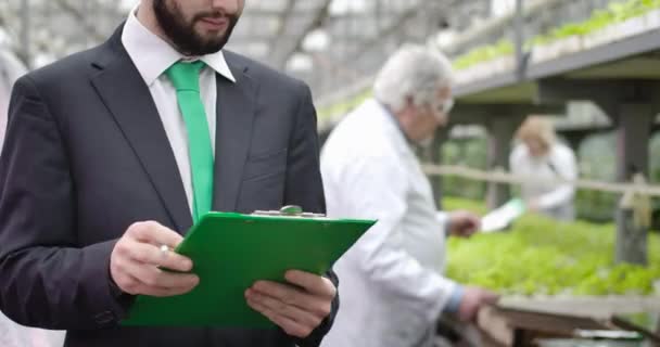 Tanımlanamayan genç bir beyaz adam arka planda çalışan olgun bir tarım uzmanına dönüşüyor ve belgelerde sebzelerin büyümesi hakkında veriler yazıyor. Organik gıda üretiminin kontrolü. Sinema 4k ProRes Merkezi — Stok video