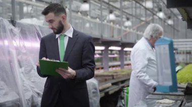 Genç Kafkasyalı müfettişten, arka planda bitkilerle çalışan olgun biyologlara notlar yazma konusunda odak değişiklikleri. Glasshouse 'daki organik gıda üretimini kontrol eden yakışıklı bir denetçi. Tarım.