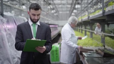 Genç yakışıklı beyaz erkek denetçinin portresi kağıtlardaki bitkilerin büyümesi hakkında veri yazıyor ve bilgi vermek için kıdemli profesyonel tarım uzmanına başvuruyor. Üretim kontrolü, tarım.