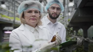 Şişesinde gübre olan yaşlı beyaz bir kadının ve cam evde poz veren yakışıklı bir adamın yakın plan portresi. Profesyonel tarım uzmanları serada organik sebzelerle ilgileniyor..