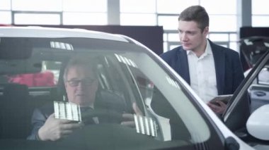 Kendine güvenen genç beyaz araba satıcısı aracın içinde oturan yaşlı müşteriye danışmanlık yapıyor. Başarılı bir yetişkin iş adamı araba galerisinde otomobil satın alıyor. İş, başarı, endüstri.