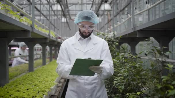 Young confident Caucasian man in protective workwear writing down, looking at camera and smiling. Camera approaching to professional agronomist working in glasshouse. Agriculture, cultivation, job. — Stock Video