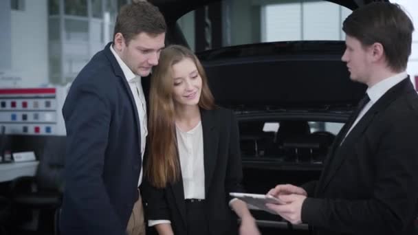 Brunett Kaukasiska langare visar bil interiörer för unga par. Handlare reklam fordon till framgångsrika kunder. Bilhandlare, showroom, bilindustri. — Stockvideo