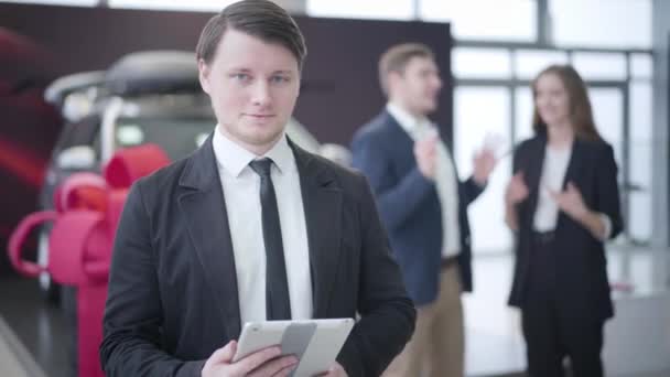 Joven morena hombre caucásico con los ojos azules mirando a la cámara, sacudiendo la cabeza y sonriendo como pareja de compradores abrazando al fondo. Distribuidor de venta de vehículos en concesionario de automóviles. Empresas, industria . — Vídeos de Stock