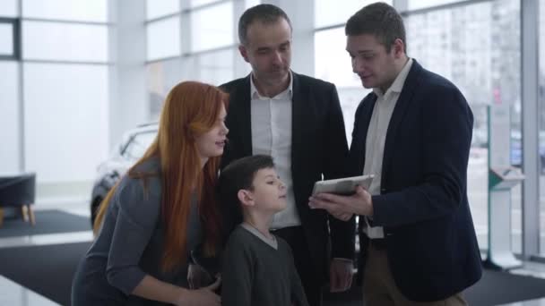 Família caucasiana conversando com revendedor de carros no showroom. Marido, esposa e menino escolhendo novo veículo na concessionária. Mãe criação filho e mostrando-lhe traficantes tablet. Indústria automobilística, compra . — Vídeo de Stock