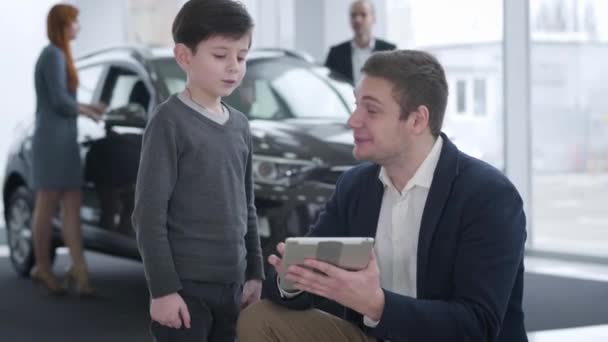 Портрет позитивного молодого кавказького торговця автомобілями, який розважає маленького хлопчина брюнетки, а його батьки вибирають новий автомобіль на задньому плані. Торгівля, бізнес, служба.. — стокове відео