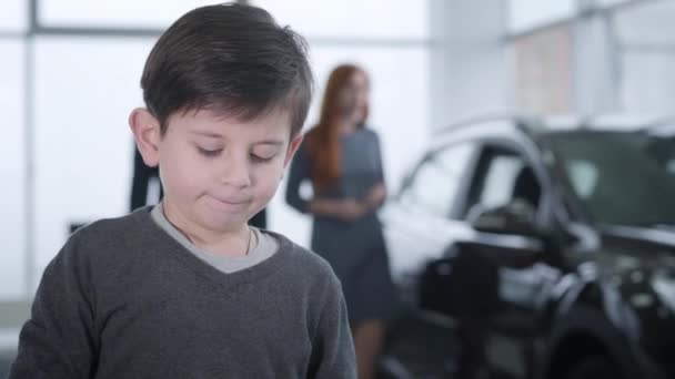 Close-up de menino morena branca bonito segurando as chaves do carro e olhando para os pais olhando para o novo veículo. Um rapazinho a comprar automóveis com o pai e a mãe na concessionária. Estilo de vida, família . — Vídeo de Stock