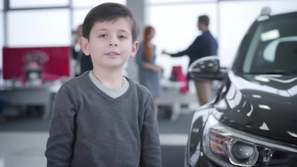 Portrait de heureux petit garçon caucasien montrant pouce vers le haut dans la concession automobile. Les parents font un marché en arrière-plan. Mode de vie, joie, bonheur, succès . — Video
