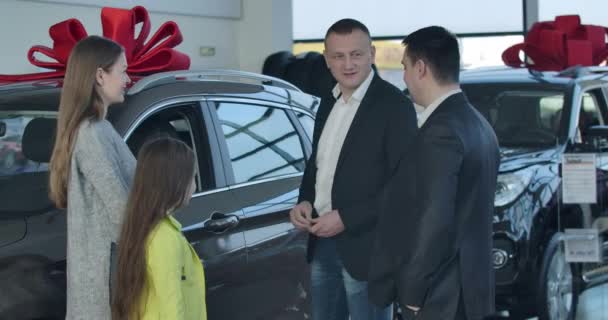 Θετική Καυκάσιος έμπορος δίνει τα κλειδιά του αυτοκινήτου στον άνθρωπο, χειραψία πελατών. Χαρούμενο κορίτσι που δείχνει τη χειρονομία νίκης στην κάμερα. Ευτυχισμένη οικογένεια που αγοράζει αυτοκίνητα στην αντιπροσωπεία. Κινηματογράφος 4k ProRes HQ. — Αρχείο Βίντεο