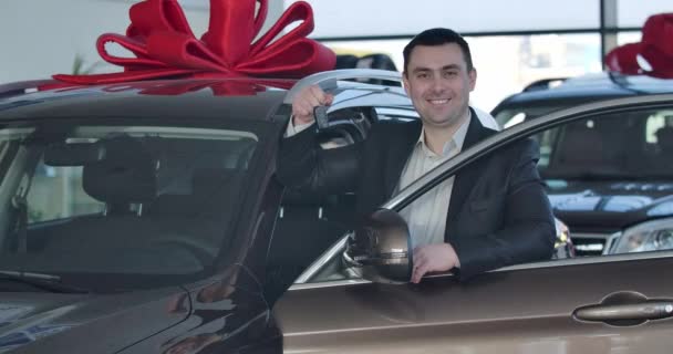 Retrato de un joven caucásico parado en un coche nuevo y alardeando de llaves. Feliz morena hombre de negocios comprando vehículo en concesionario. Propietario del auto posando en la sala de exposición. Sede del cine 4k ProRes . — Vídeos de Stock