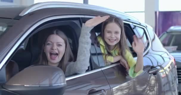 Pozytywna kaukaska matka i córka machające z nowego eleganckiego czarnego samochodu w salonie. Uśmiechnięta szczęśliwa kobieta i mała dziewczynka ciesząca się zakupem pojazdu w salonie. Kino 4k ProRes kwatera główna. — Wideo stockowe