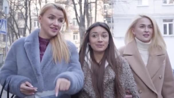 Detailní portrét tří okouzlujících mladých žen, které se procházejí po ulici a povídají si. Stylové bělošky v podzimních kabátech procházející se venku a odcházející. Zpomalený pohyb. — Stock video