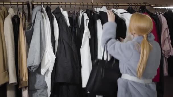 Stijlvolle jonge blanke vrouw praat zwarte jurk van hanger in de winkel. Rijk stijlvol meisje winkelen in de mode boetiek en glimlachen. Lifestyle, shopaholisme. Langzame beweging. — Stockvideo
