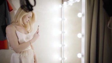 Soyunma odasında fotoğraf çeken şık, beyaz bir kadının yan görüntüsü. Zarif bej elbiseli, akıllı telefon tutan ve moda butiğinde aynaya bakan sarışın zengin bir kadın. Yavaşla.
