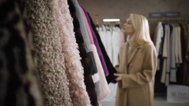 Die Kamera nähert sich einer jungen blonden Kaukasierin, die sich für einen Wintermantel in Boutique entscheidet. Selbstbewusste elegante Dame beim Einkaufen. Konsum, Lifestyle, Mode. Zeitlupe. — Stockvideo